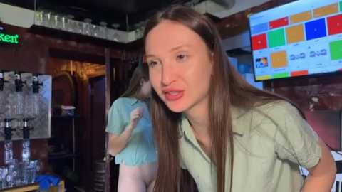 Video of a woman with long brown hair, wearing a light green shirt, standing in a bar with a colorful digital screen and bar equipment in the background.