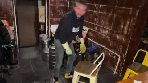 A video of a man in a black hoodie, yellow gloves, and jeans, leaning on a wooden stool in a dimly lit, cluttered room with a brown brick wall and beer kegs.
