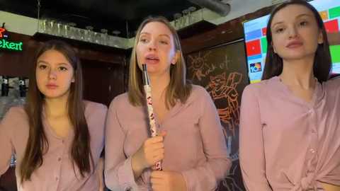 Video of three young women in light pink button-up shirts, one holding a fork and knife, in a dimly lit bar with a colorful TV screen and bar counter in the background.