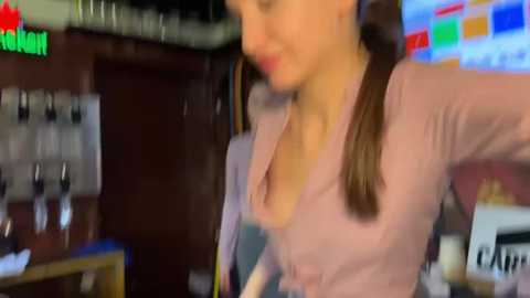 A blurred video of a woman with a ponytail in a bar, wearing a pink blouse, standing in front of a TV displaying sports news.