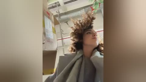 Video of a woman with voluminous, curly brown hair, wearing a light grey robe, standing in a dimly lit, industrial room with white walls, exposed pipes, and a red fire extinguisher.