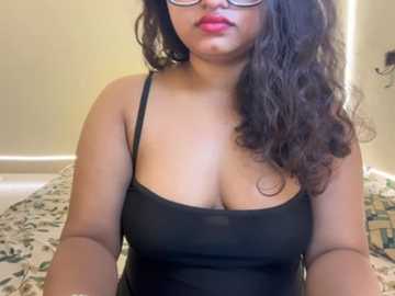 Video of a South Asian woman with medium-dark skin, long curly black hair, and glasses, wearing a black spaghetti-strap top that reveals cleavage, sitting on a bed with floral sheets.