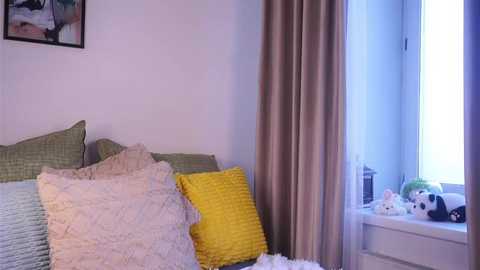 Video of a cozy bedroom with a bed covered in light green and white bedding, a yellow pillow, and beige curtains. A window with a white frame holds stuffed toys.