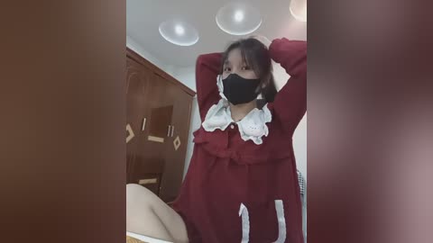 Video of an East Asian woman with medium skin tone, dark hair, and wearing a red dress with white ruffles, black face mask, and arms raised, standing in a dimly lit room with wooden cabinets and a bed in the background.