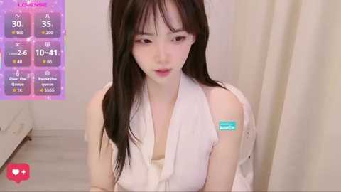 Video of a pale-skinned, slender Asian woman with long black hair, wearing a white sleeveless top, sitting on a bed with beige curtains. A \"LIVE\" overlay displays her stats, including 50% health and 59% stamina.
