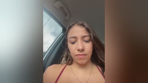 A video of a young Latina woman with long brown hair, wearing a red spaghetti strap top, sitting in a car, looking introspective, with a blurred background.