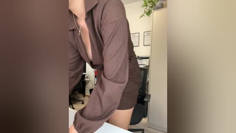 Video of a woman in a brown shirt and shorts bending over a table in an office, with potted plants and framed certificates in the background.