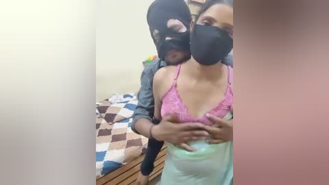 Video: A woman in a pink lace bra and light green skirt is held by a man in a black mask, standing in a bedroom with a patterned bedspread and a wooden dresser.