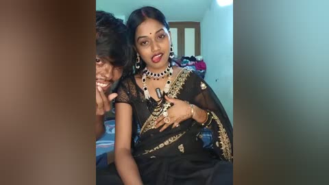 A video of a South Asian woman in a black sari with intricate gold embroidery, wearing heavy jewelry, and a man with a beard, both smiling, in a dimly lit room with a bed and clothes in the background.