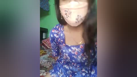 Video of a woman in a blue floral dress, with a face mask and black tape over her mouth, standing in a cluttered, brightly lit room with green walls.