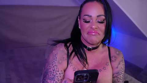 Video of a tattooed woman with medium skin tone, black hair, and large breasts, wearing a black choker and leopard-print top, taking a selfie indoors under purple lighting.