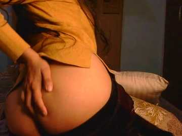 Video of a woman's lower back, wearing a mustard-yellow jacket and black panties, posing seductively on a bed with floral-patterned sheets, in a dimly lit room.