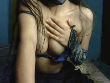 Video of a woman with light skin, long brown hair, wearing a blue lace bra, holding her breasts. She is in a dimly lit room with a blue wall and a dark floor.