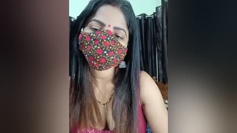 A video of a South Asian woman with long dark hair, wearing a colorful floral face mask and a pink blouse. She has a red dot on her forehead.