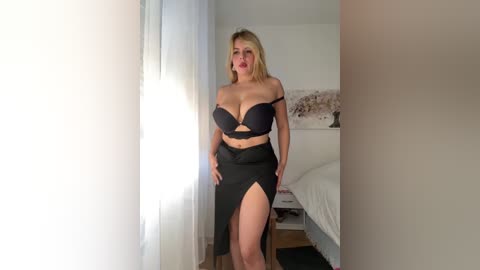 Video of a blonde Caucasian woman in black lingerie, standing confidently in a bright, modern bedroom with white bedding and sheer curtains.