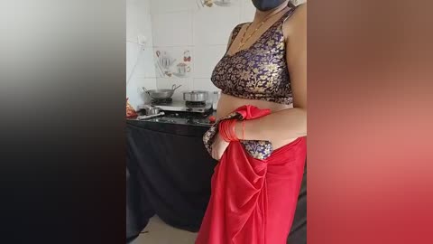 Video of a person in traditional Indian attire with a floral blouse, red skirt, and a headscarf, standing in a kitchen with white tiled walls and a black countertop.