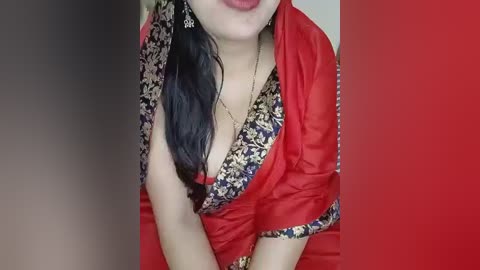 Video of a woman with fair skin, wearing a red sari with a floral pattern and a black blouse. She has long, dark hair and is indoors against a red background.