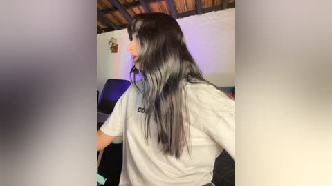 A video of a young woman with long black hair, wearing a grey t-shirt, standing in a dimly lit room with a wooden ceiling and white walls.