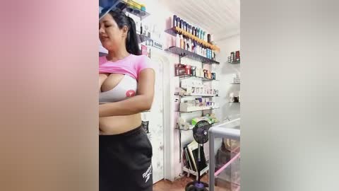 Video of a Latina woman with medium skin tone and large breasts, lifting a pink shirt to reveal a white bra, in a brightly lit beauty salon with shelves of products and a hairdryer.