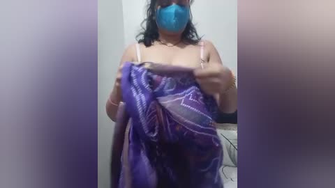 A video of a woman with a blue face mask, purple and white patterned cloth covering her breasts, and medium brown skin, taken indoors with a blurred background.