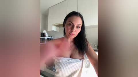 A video of a young woman with long black hair, fair skin, and a slender physique, wearing a white, off-the-shoulder towel, standing in a modern, minimalist kitchen with beige cabinets and stainless steel appliances.