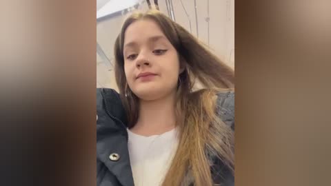 Video of a young woman with long, straight brown hair, wearing a black jacket and white top, smiling. Background features a blurred, beige wall.