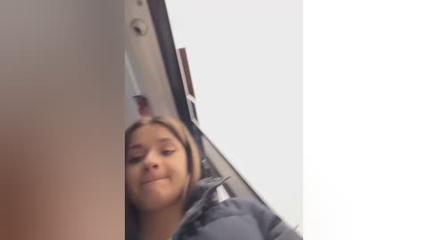 A video shows a young woman with light brown skin and straight, shoulder-length hair, wearing a dark jacket, taking a selfie in a dimly lit room with a white wall and a brown door.