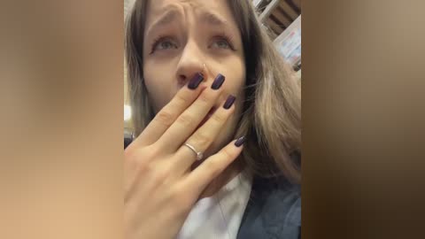 Video of a woman with long brown hair and blue eyes, covering her mouth with a hand. She has dark purple nail polish and wears a silver ring. Background shows a blurred library with shelves.