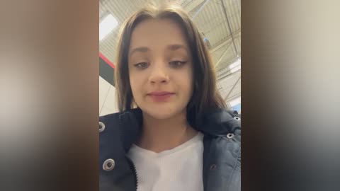 Video of a young girl with straight, medium-length brown hair, fair skin, and a light complexion, wearing a white t-shirt under a blue puffer jacket. The background features a modern, industrial space with exposed pipes and a high ceiling.