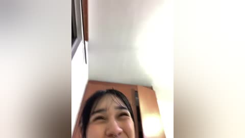 Video of a young Asian woman with long black hair, smiling, captured from a low angle, showing her chin and upper chest. Background includes a wooden wall and a bright light source.