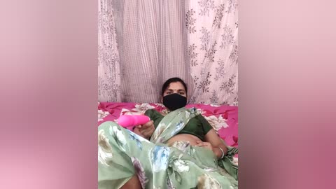 A video of a woman lying on a bed with a floral green sari, wearing a black face mask, holding a pink vibrator, set against pastel curtains and a pink background.