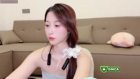 Video of an East Asian woman with long, straight brown hair, wearing a light blue strapless dress, sitting on a beige sectional couch with a beige cushion and a plush, beige stuffed animal. She wears a white, flower-patterned scrunchie.