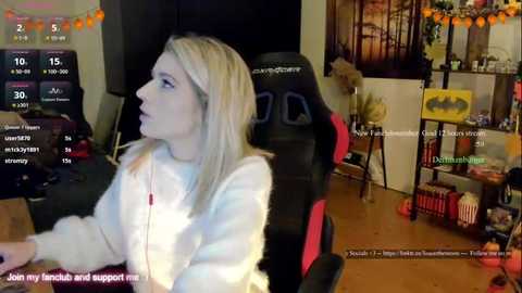 Video of a young woman in a gaming chair, wearing a white hoodie, playing a video game. Background shows a cluttered room with a Batman poster and a shelf.