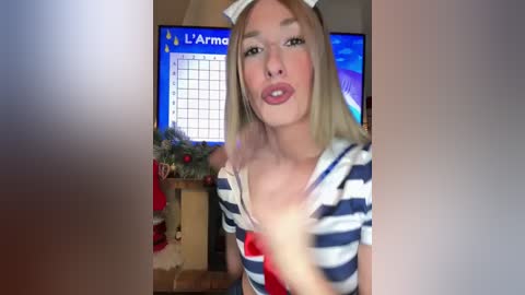 Video of a young blonde woman in a striped shirt, blowing a kiss with a Santa hat, in a cozy, festive living room with Christmas decorations and a TV screen displaying \"La Armada.\