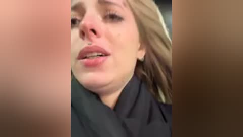 A video of a distressed, tearful young woman with blonde hair, fair skin, and a black top. Her face is partially obscured by another person's hand. The background is blurred.