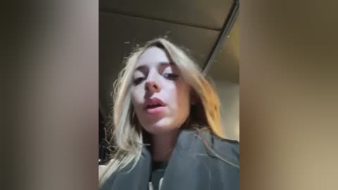 Video of a blonde woman with a fair complexion, wearing a black jacket, taken from a low angle, indoors with a ceiling light visible.