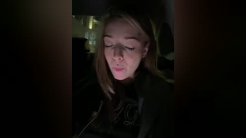 Video of a blonde woman with fair skin, wearing a black jacket, looking intently at a laptop screen in a dimly lit room, with blurred background objects.