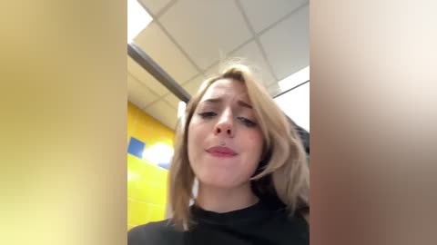 Video of a young, blonde woman with fair skin, wearing a black shirt, standing in a brightly lit, modern restroom with yellow and blue tiled walls.