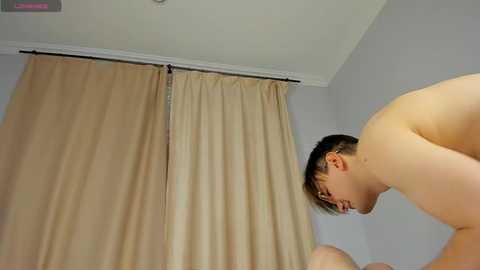 Video of a naked Asian man with short dark hair, wearing glasses, leaning forward with his face close to beige curtains in a minimalistic room with white walls.