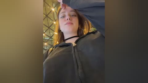 Video of a young Caucasian woman with light skin, long straight brown hair, and a slender physique, wearing a black choker and dark jacket. She stands against a background of geometric metal panels with warm, glowing fairy lights.