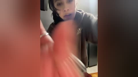A blurry video of a young Asian woman with long black hair, wearing a grey hoodie, looking surprised as she takes a selfie through a window.