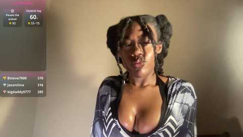 Video of a Black woman with medium-dark skin, large breasts, and two pigtails. She wears a low-cut, black and white patterned dress, and her expression is neutral. Background is a plain beige wall.