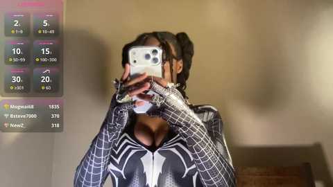 Video of a person wearing a black and white Spider-Man suit, holding a phone to their face, against a beige wall.