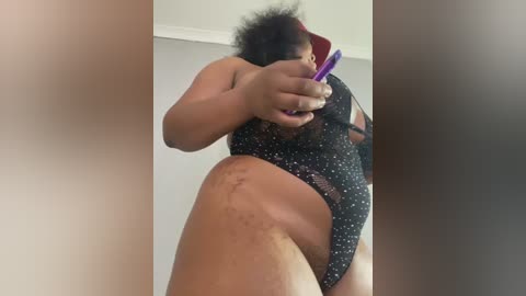 Video of a plus-sized Black woman with curly hair in a black polka-dotted bodysuit, holding a purple vibrator, taken from a low angle.