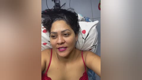 A video of a woman with dark hair, wearing a red lace bra, lying on a bed with floral-patterned pillows, smiling.