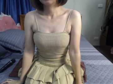 Video of a slender East Asian woman with short brown hair, wearing a beige ribbed tank dress with thin straps, sitting on a bed with blue and white striped sheets, in a simple bedroom with a wooden headboard and blue curtains.