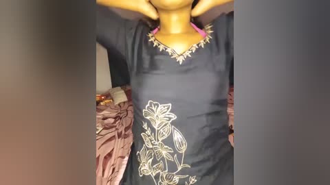 Video of a woman in a dark, embroidered, long-sleeved shirt with floral designs and a pink choker. She has her hands behind her head, standing in a dimly lit room with a brown fabric draped on her left side.