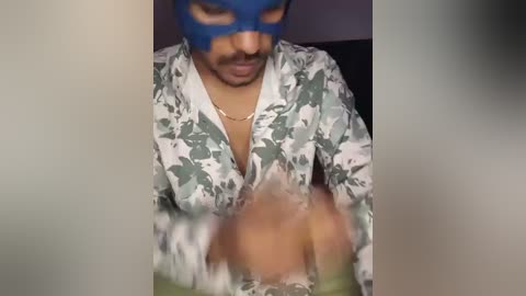 Video of a man with medium skin tone and trimmed beard wearing a blue mask and a floral shirt, partially unbuttoned, revealing a glimpse of his bare chest.