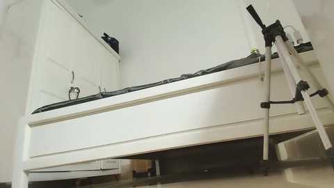 Video of a white, unmade twin bed in a minimalist room, with a white dresser and a black cat sitting on top. The floor is light beige tile.