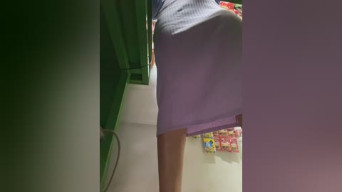A video captures a woman's lower body, wearing a tight, purple mini dress, standing in a supermarket aisle. Green shelves and colorful snacks are visible in the background.